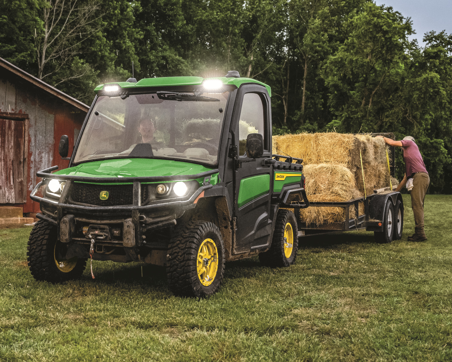 A John Deere Gator with a covered cab pulls a trailer with square hay bales.
