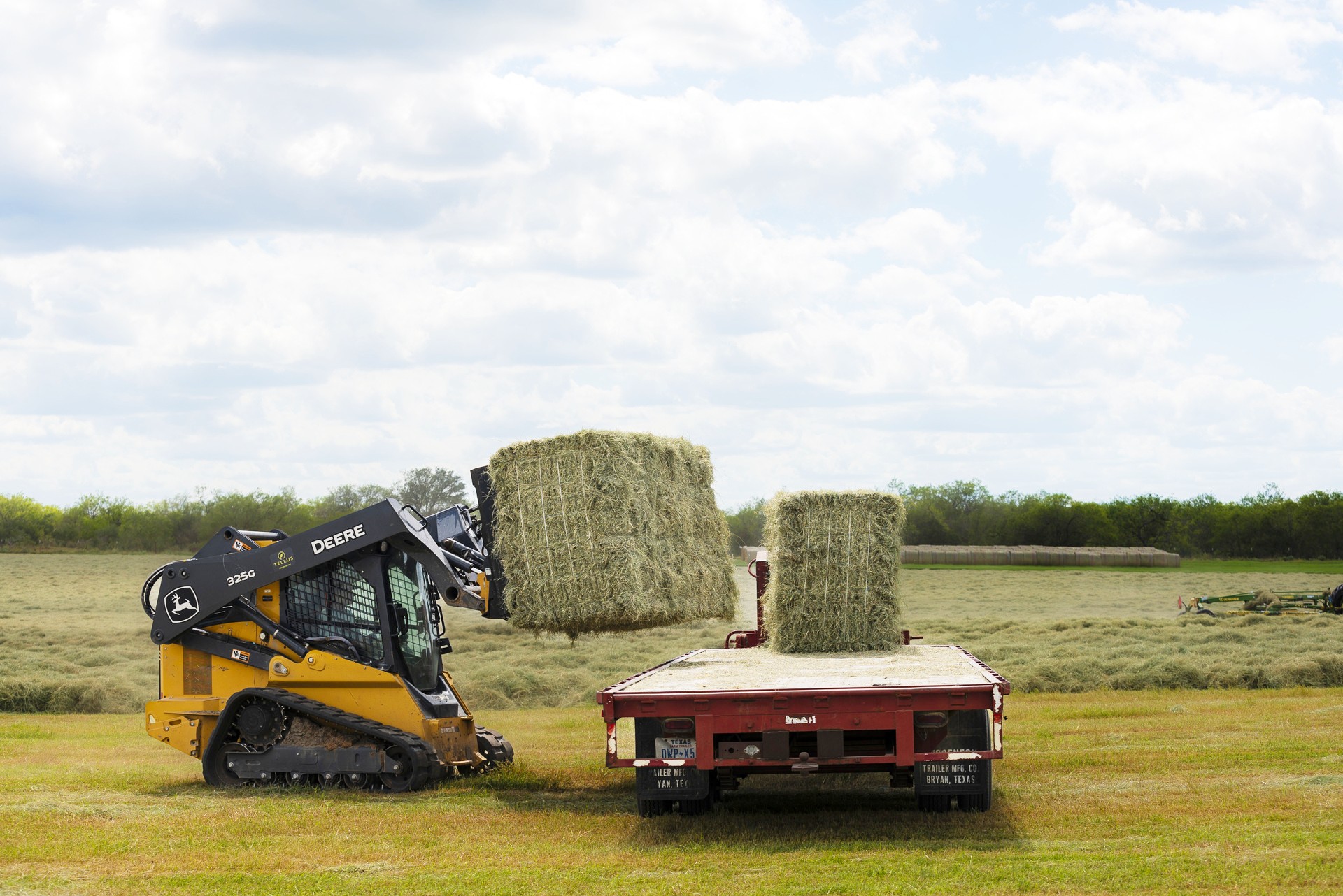 A compact track loader lifts a square hay bale onto a trailer in Texas.