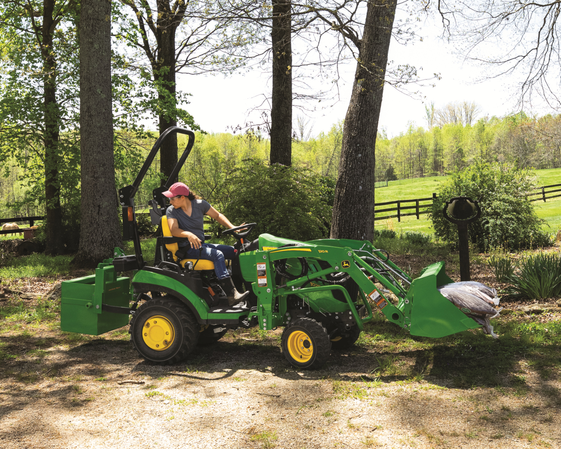 A John Deere tractor with a front-end loader and box blade attachment clear land in the fall.