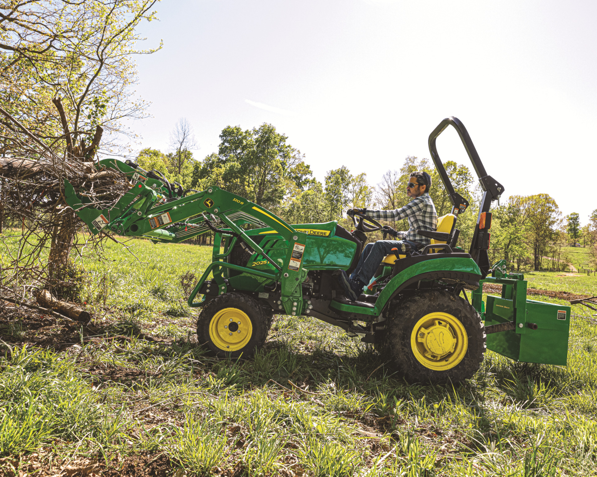 A homeowner uses a John Deere tractor with a front-end loader to clear brush on their land.