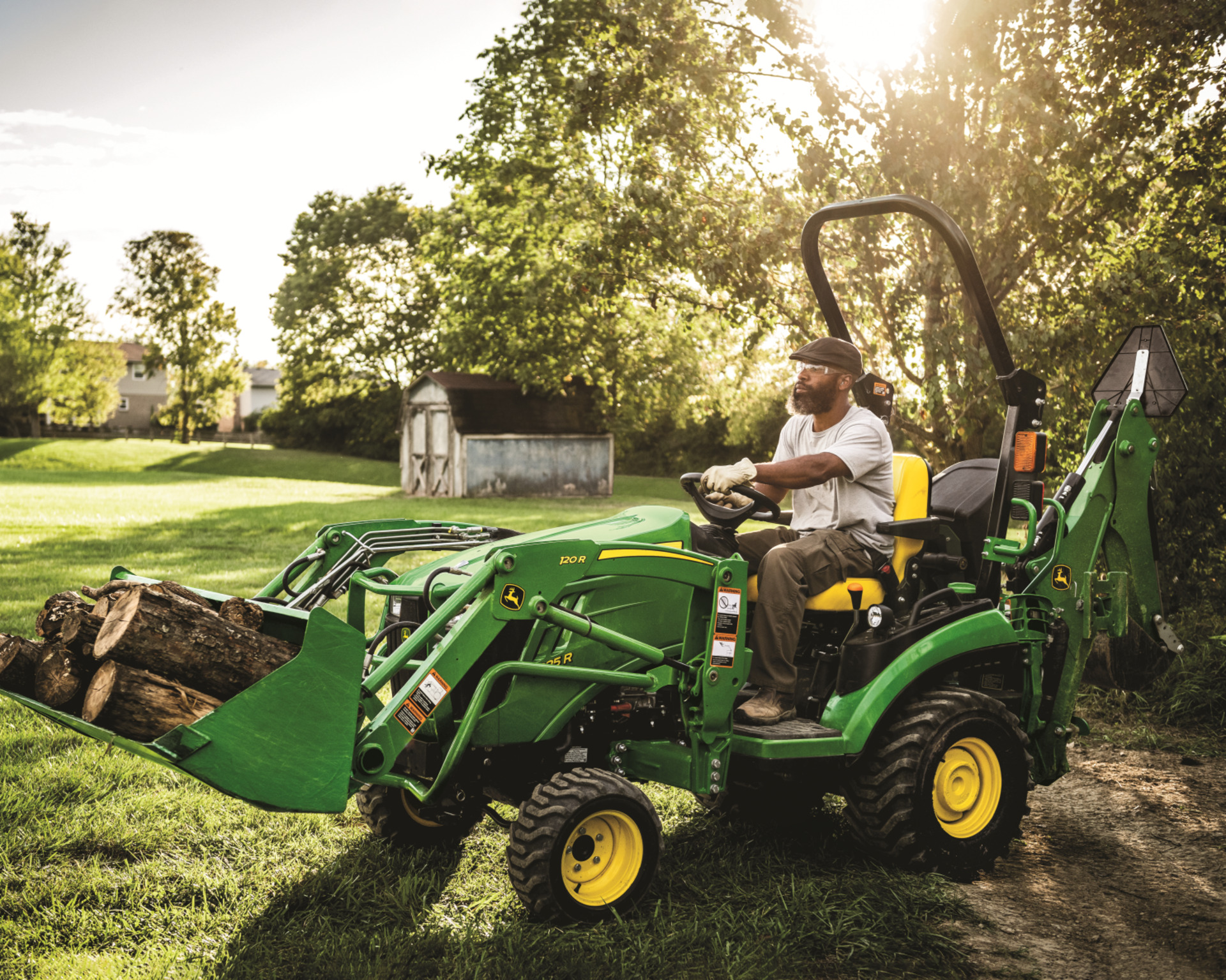 A homeowner uses a John Deere compact tractor to move logs on their property with a front-end loader